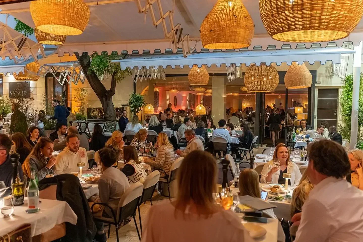Busy outdoor dining at La Mome restaurant in Cannes with rattan lamps and guests.